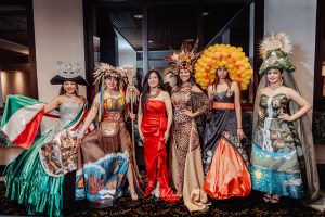 Group portrait of seven women on stage wearing elaborate costumes including traditional embroidered Mexican dresses, ornate headdresses with floral and decorative elements, all in formal attire