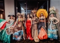 Group portrait of seven women on stage wearing elaborate costumes including traditional embroidered Mexican dresses, ornate headdresses with floral and decorative elements, all in formal attire