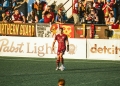 Soccer player in a red team kit controlling a ball on the pitch during a match, with a crowded stadium section behind featuring fans in various team colors and a large banner reading "Look what I found in the dumpster!" with a cartoon character, plus signage for Hartford and Pabst Light along the sideline.