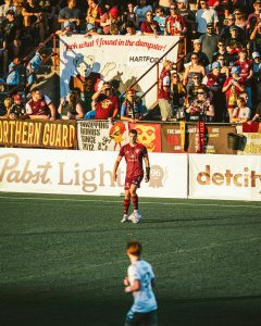 Soccer player in a red team kit controlling a ball on the pitch during a match, with a crowded stadium section behind featuring fans in various team colors and a large banner reading "Look what I found in the dumpster!" with a cartoon character, plus signage for Hartford and Pabst Light along the sideline.