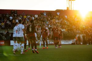 Soccer match at sunset with players in white and dark red uniforms on the field. A crowded stadium is visible in the background with golden hour lighting illuminating the scene