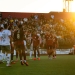 Soccer match at sunset with players in white and dark red uniforms on the field. A crowded stadium is visible in the background with golden hour lighting illuminating the scene