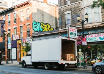 Urban street scene showing brick buildings with colorful graffiti, a white delivery truck, storefronts including an "International Fish Market" with pink flowers displayed, and street-level signage.