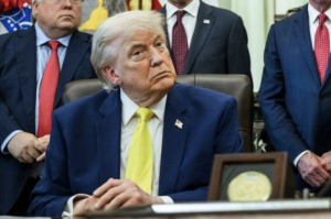 Man in a dark blue suit and yellow tie seated at an official desk surrounded by other men in business suits in what appears to be a formal government or administrative office setting.