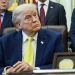Man in a dark blue suit and yellow tie seated at an official desk surrounded by other men in business suits in what appears to be a formal government or administrative office setting.